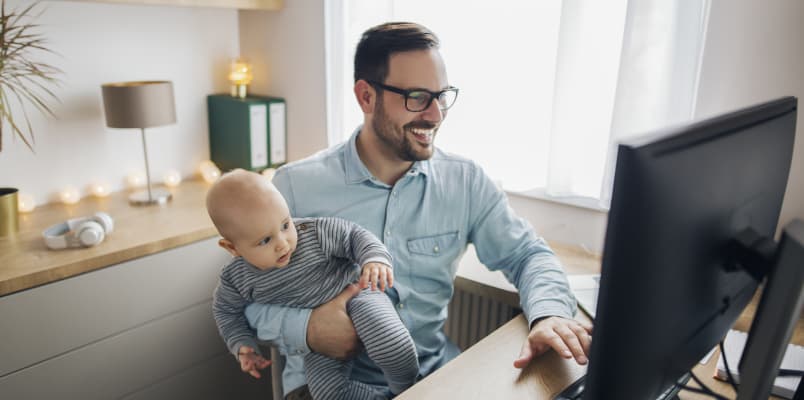A grinning man with an infant in his arms while working on a computer.