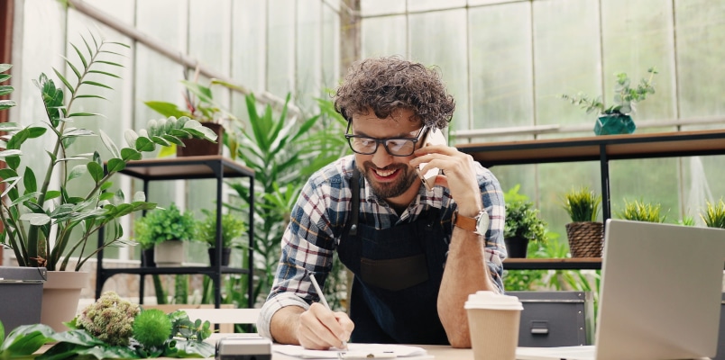 A smiling man wearing an apron working in a greenhouse, talking on the phone and taking notes at a desk with a laptop while reviewing his small business tax deductions.