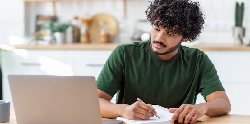 Person at a kitchen table reviewing notes on a laptop while writing in a notebook.
