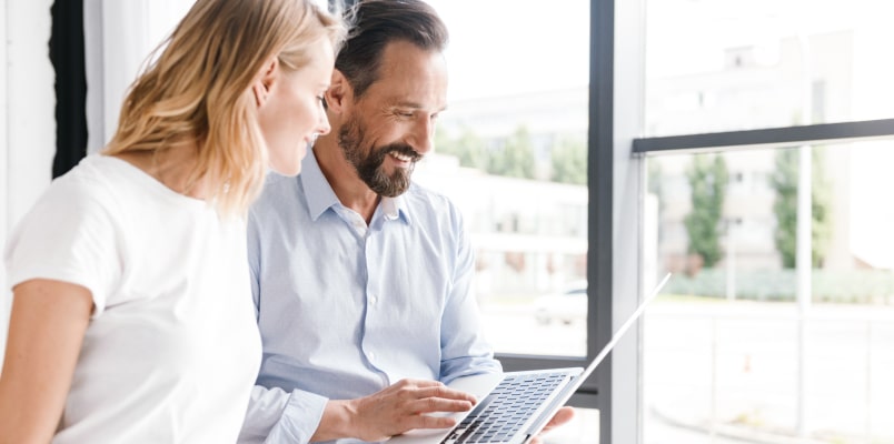 A smiling man and woman sitting by a bright window, looking at a laptop together while discussing 401k tax benefits and retirement planning.