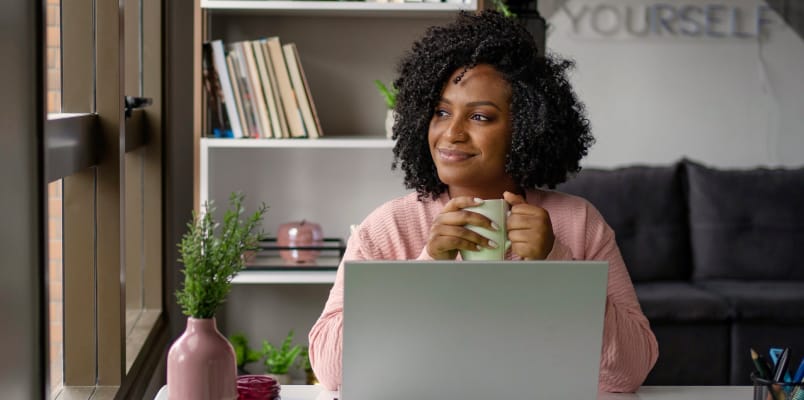 A joyful young African American woman with long curly hair, sitting in front of her laptop and enjoying her coffee.