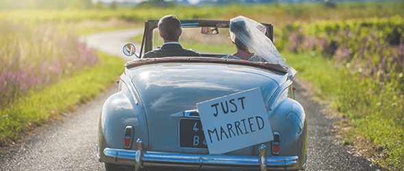 A bride and groom drive in a blue convertible with a sign reading 