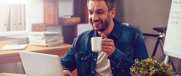 A smiling man holding a cup of coffee in one hand and typing on laptop with the other hand
