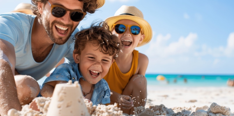 A dad and his two young children smiling and laughing as they build a sandcastle on a beach in the summer