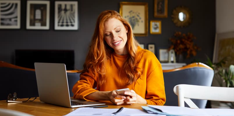 A red-haired woman in an orange sweater at her work desk checking out her cellphone.