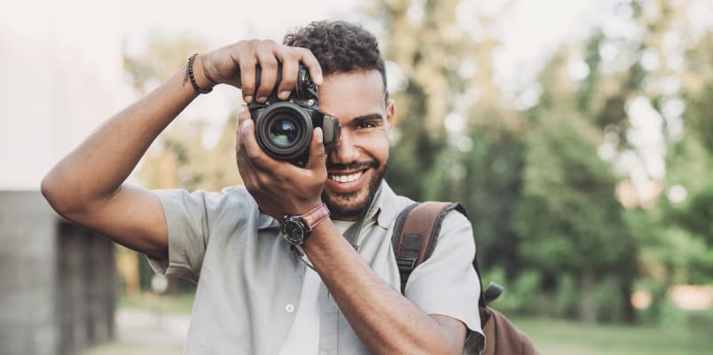 A smiling man looks forward while looking through a camera lens held up to his right eye.