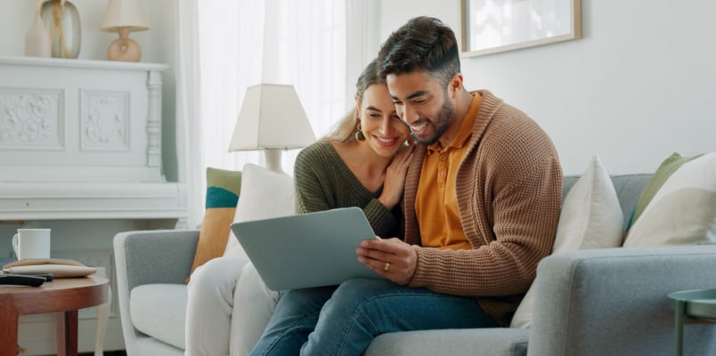 A man and woman wearing cozy sweaters sitting on a couch and smiling at a tablet