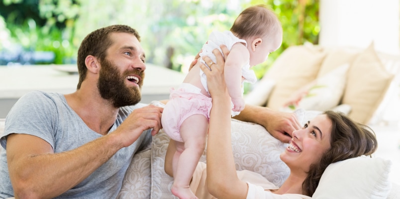A smiling man sits next to a smiling woman who is lying on a couch holding her baby above her head