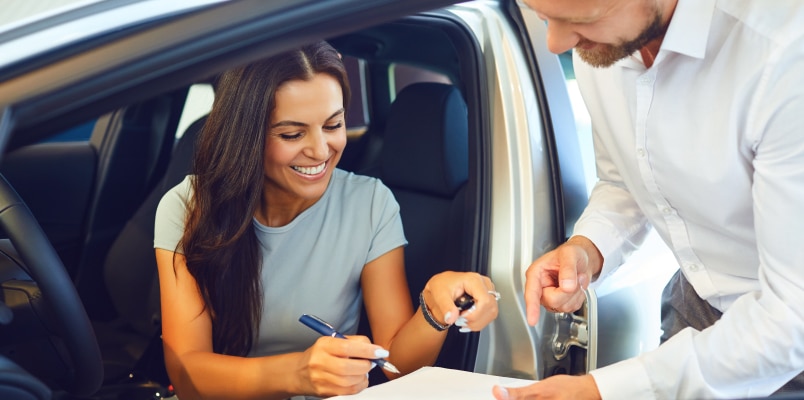 A young woman business owner sitting in a new vehicle and signing the purchase paperwork held by a car salesman