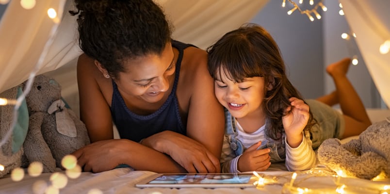 A woman and her daughter look at a tablet while lying on a bed under a sheet tent with decorative lights and stuffed animals.