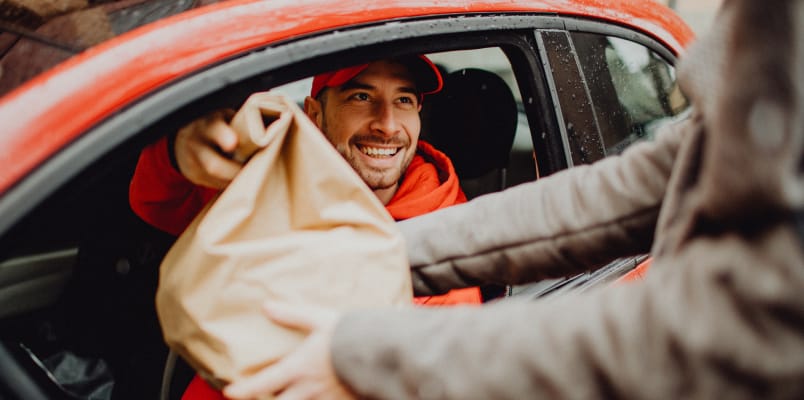 A smiling delivery driver earning side income hands a customer a bag out of his car window