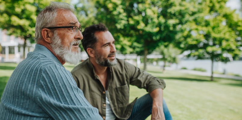 An older man and a middle-aged man sitting together outside on a sunny day, talking about financial planning and tax deductions for seniors.