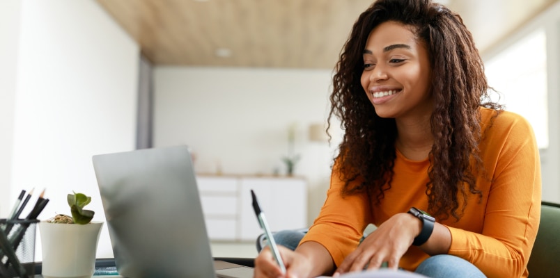 A smiling woman sitting at a desk with a laptop, taking notes while learning about the process for a nonresident filing U.S. taxes.