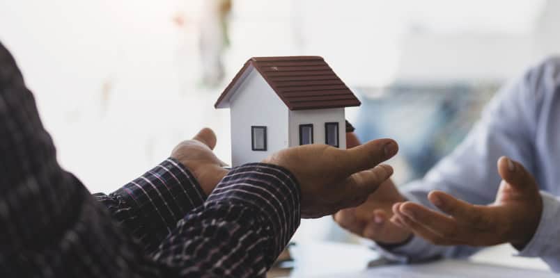 A closeup of the hands of two people across from each other at a table. One person holds out a small model house for the other person to receive.