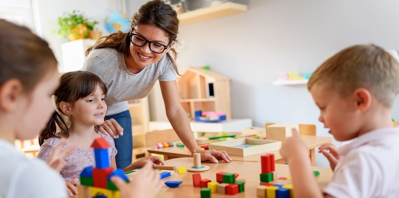 A woman supervises young children playing with blocks in a childcare setting illustrating expenses reported on Form 2441 for the child and dependent care credit.