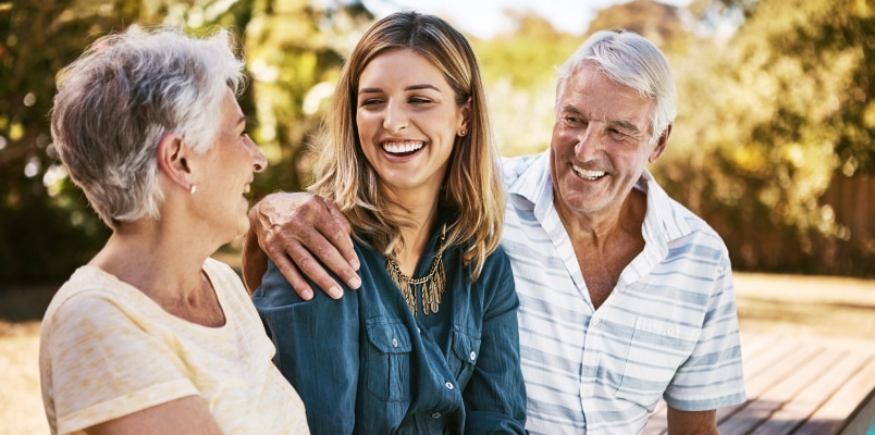 An older couple and their adult daughter sitting together outdoors, smiling and talking while discussing gift tax implications and the financial aspects of supporting adult children.