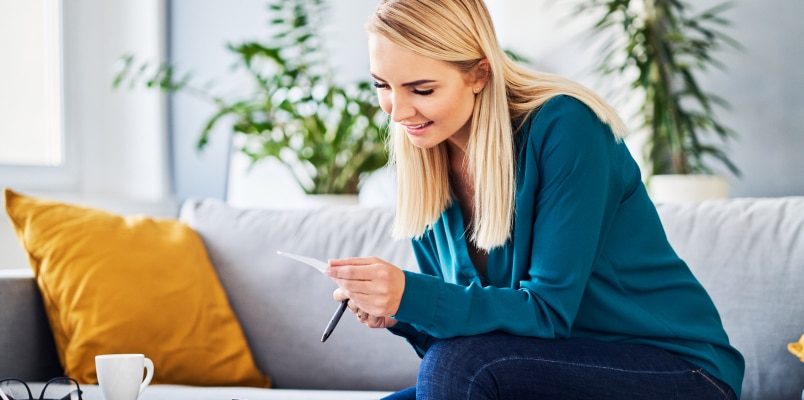 A woman sitting on a sofa with a pen in hand, smiling as she reviews a receipt while calculating itemized deductions for her tax return.