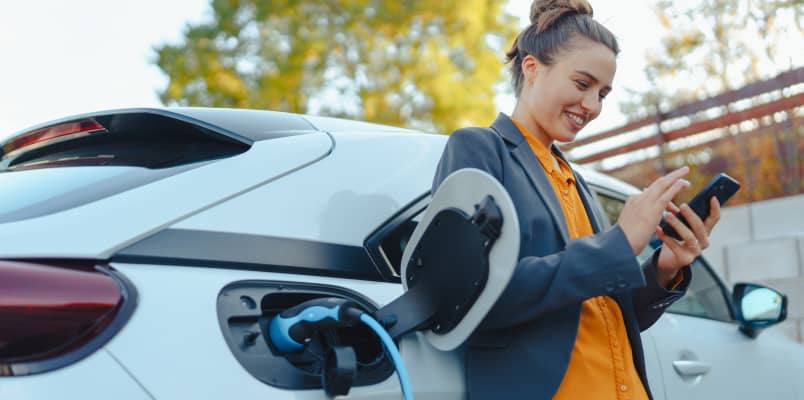 A smiling woman leans against her electric vehicle as she waits for it to charge