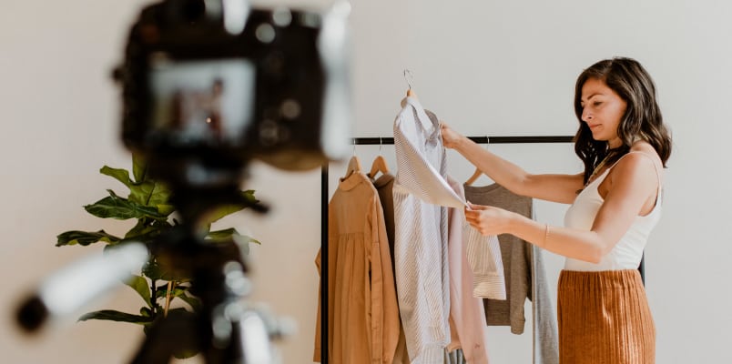 A female content creator stands before a camera while holding a shirt in front of a rack of clothes.