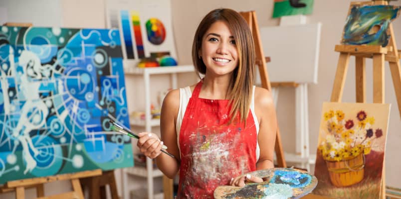 An artist smiling while holding paint brushes and colorful palette in her hands.