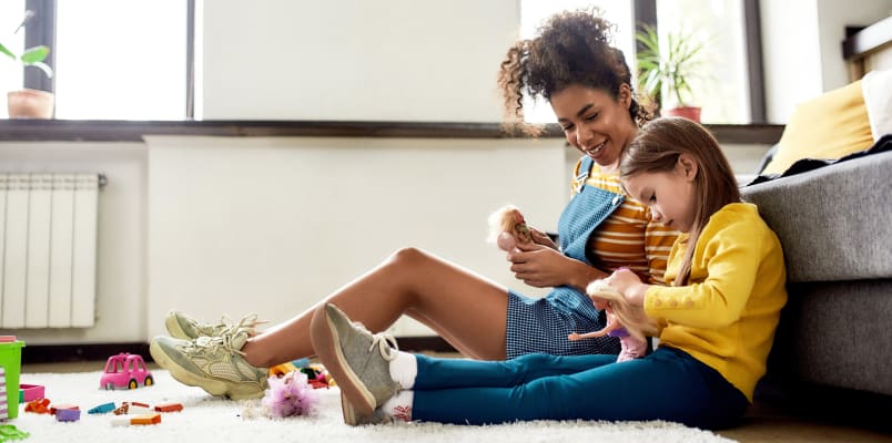 A young woman sits on the living room floor next to a little girl. They are both holding Barbie dolls.