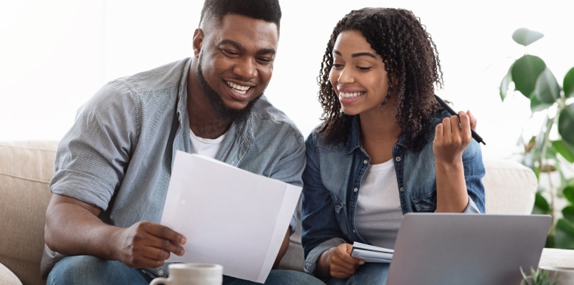 A smiling couple sitting on a couch, reviewing financial documents together in front of a laptop, illustrating a discussion about IRA vs. Roth IRA vs. 401k retirement options.