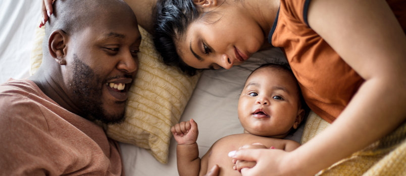 A smiling man and woman lie in bed with their grinning infant in between them.