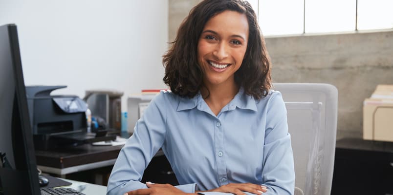 A smiling woman with brown hair and wearing a blue button-up shirt sits in an office.