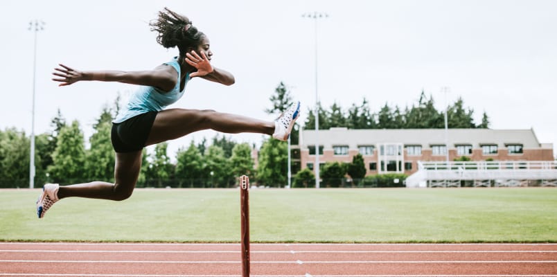 A young woman in blue running track jumps over a hurdle.