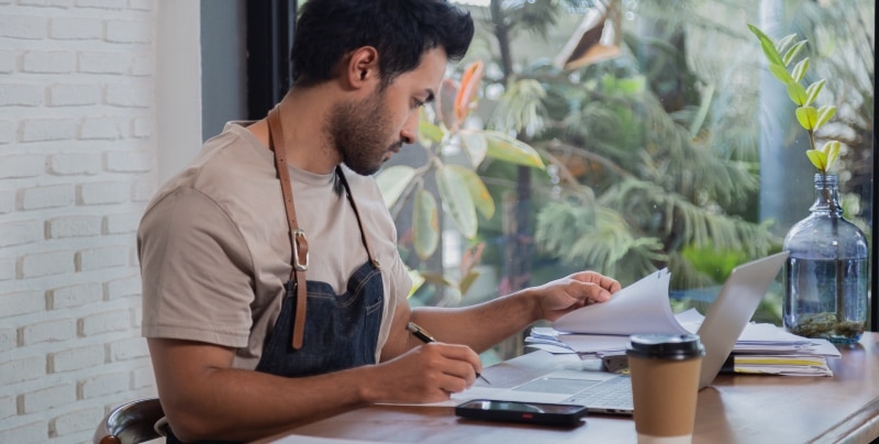 Man in barista uniform is filing taxes on laptop with a stack of paper