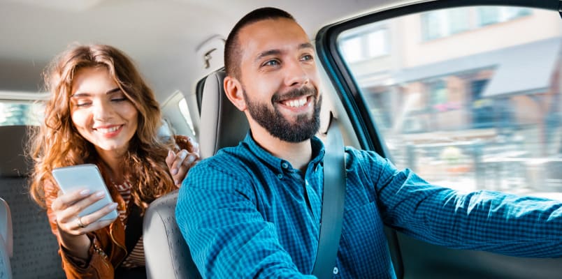A smiling man with a woman riding as a passenger in the back seat