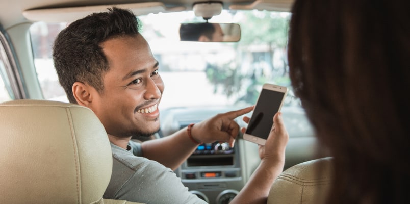 A smiling driver holding a phone and turning to look at the passenger in his backseat