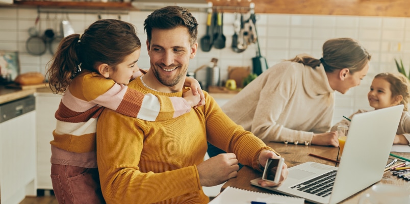 A young child smiles at her father with her arms around his neck at their kitchen island while her mother smiles at another child in the background