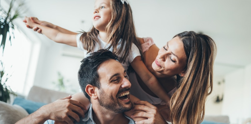 A young girl sitting on her smiling father's shoulders while her mother hugs them both