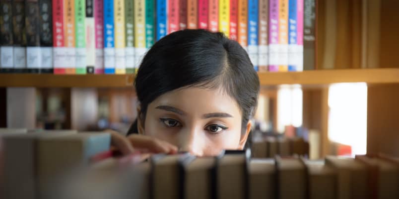 A girl standing in a library is confused about which book she should pick up for reading