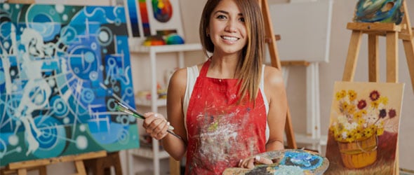 A woman smiling while holding paint brushes and palette in her hands.